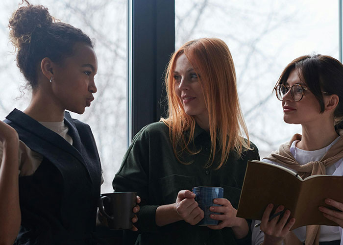 Three women discussing neurodivergent experiences while holding mugs and a book near a window in a casual setting.