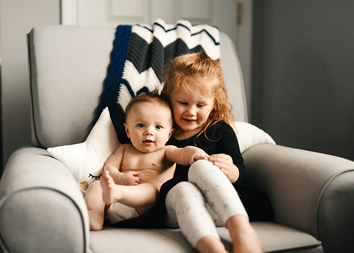 Two young children sitting on a gray armchair, highlighting the role of nannies and the internet in raising kids.