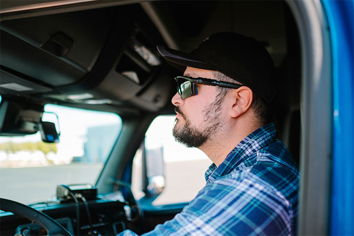 Man wearing glasses and a cap inside a truck, using theatrics for revenge on a workplace bully with scare tactics.