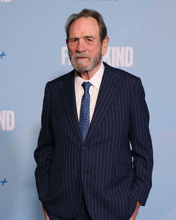 Older man with grey beard wearing a dark pinstripe suit and patterned blue tie at a media event background.