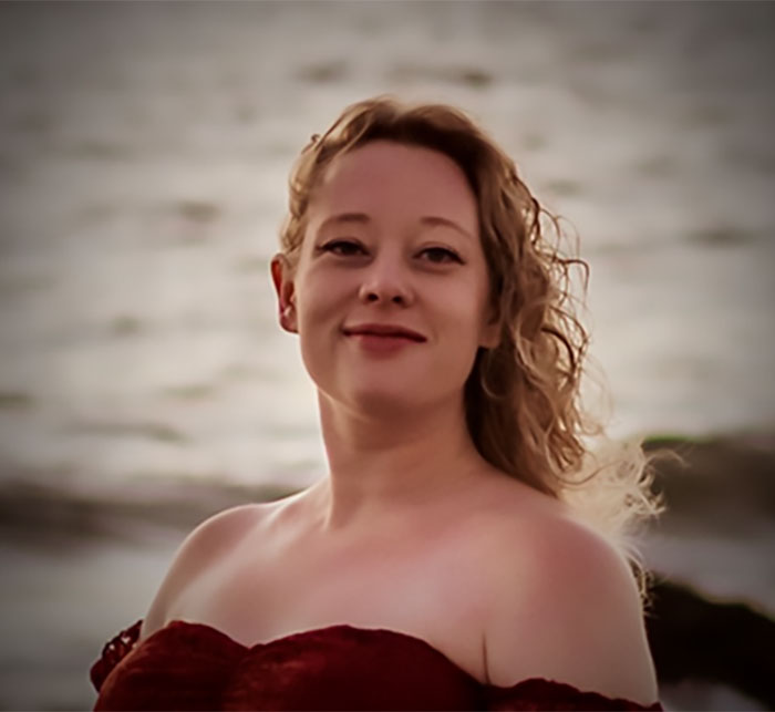 Young woman with curly hair wearing a red off-shoulder dress by the water, related to family of mom slain by ICE agent.