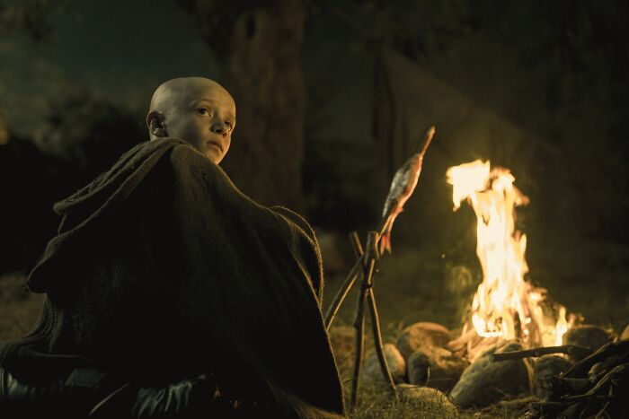 A bald character in medieval cloak sitting by a campfire with fish roasting, related to A Knight of the Seven Kingdoms.