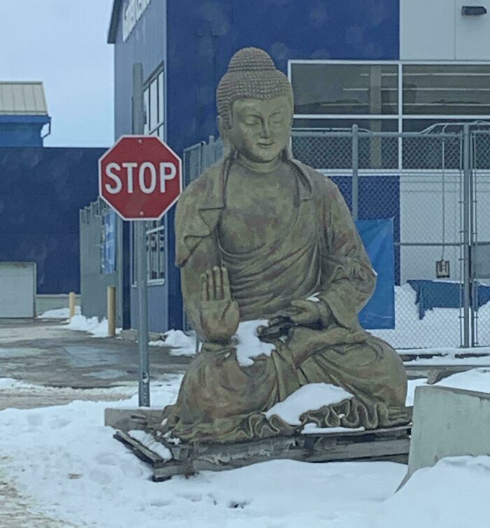 Buddha statue in snow with missing lower section next to a stop sign in an industrial area, a confusing funny picture.