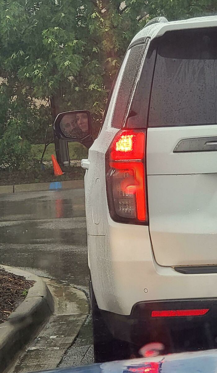 Rear view of a white SUV with a confusing reflection of a face in the side mirror on a rainy day.