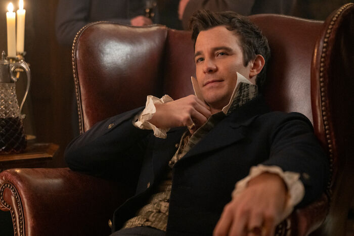 Male actor from Bridgerton Season 4 cast list seated in a leather armchair in a period costume setting