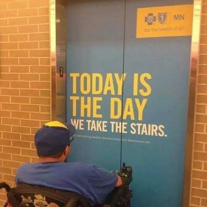 Man in wheelchair facing an elevator door with a sign about taking the stairs, reflecting dark humor memes.