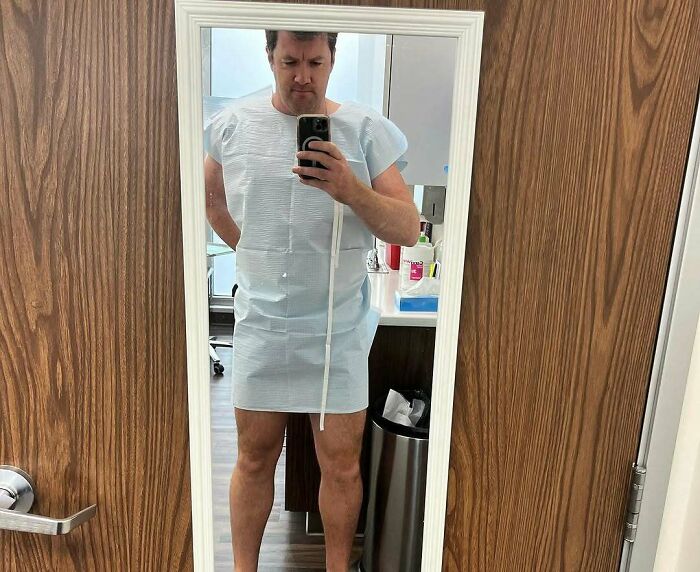 Man in a medical gown taking a mirror selfie, illustrating common struggles tall people face with clothing fit.