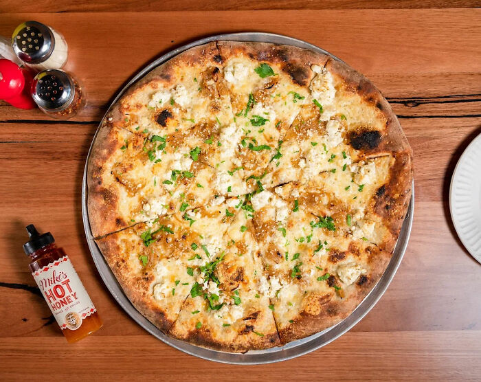 Wood-fired pizza on a metal tray with seasoning shakers and hot honey, related to pizzeria owner viral salty replies.