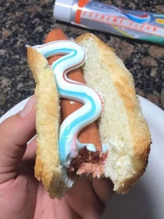 Hand holding a hot dog bun with a sausage topped with toothpaste instead of mustard in a bizarre meme image.