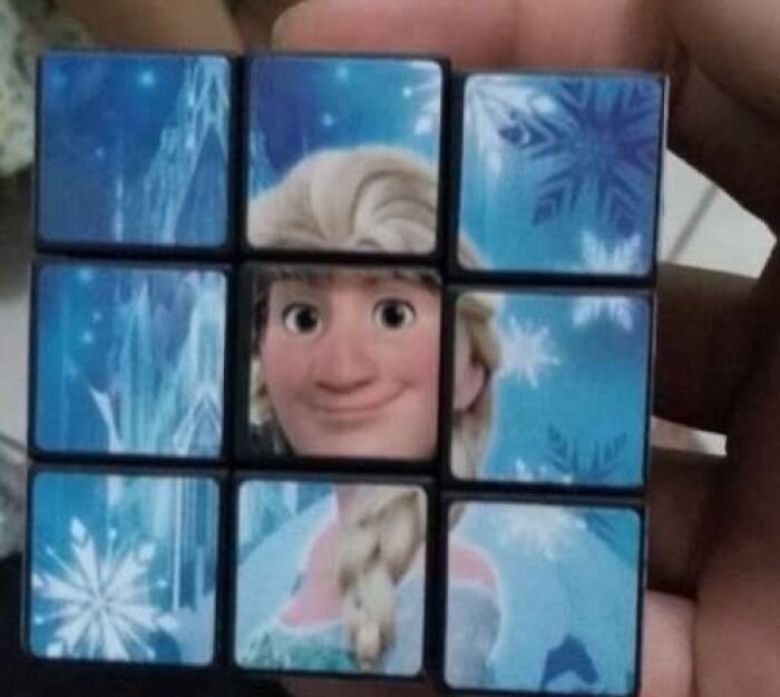 Hand holding a bizarre scrambled Rubik’s cube with a distorted cartoon character’s face and snowflake patterns.