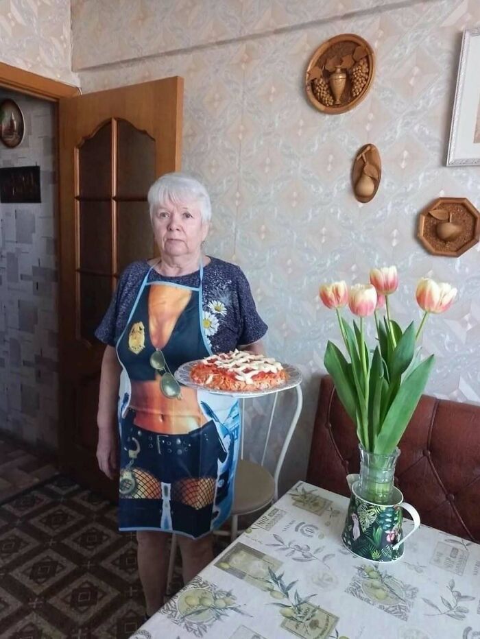Elderly woman wearing a bizarre apron holding a pizza, surrounded by home decor and a vase of tulips indoors.