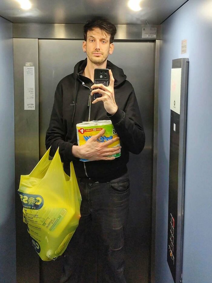 Tall person taking a mirror selfie in an elevator, holding groceries and wearing a black hoodie.