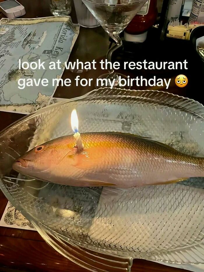 A bizarre birthday meme shows a whole fish on a plate with a lit candle stuck in it at a restaurant.