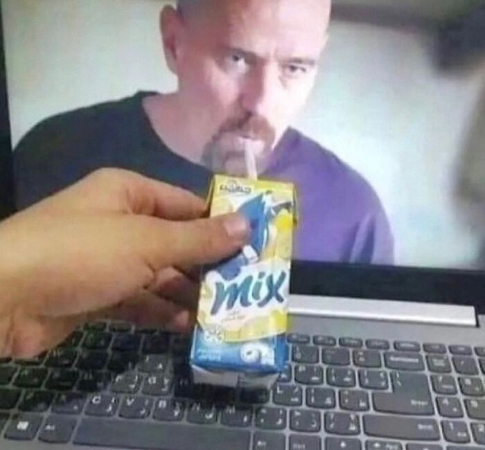 Hand holding a juice box with a straw aimed at a laptop screen showing a man drinking in a bizarre meme.