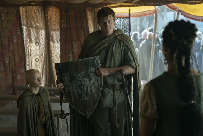 Two characters inside a tent, one holding a shield displaying house sigils from a Knight of the Seven Kingdoms.