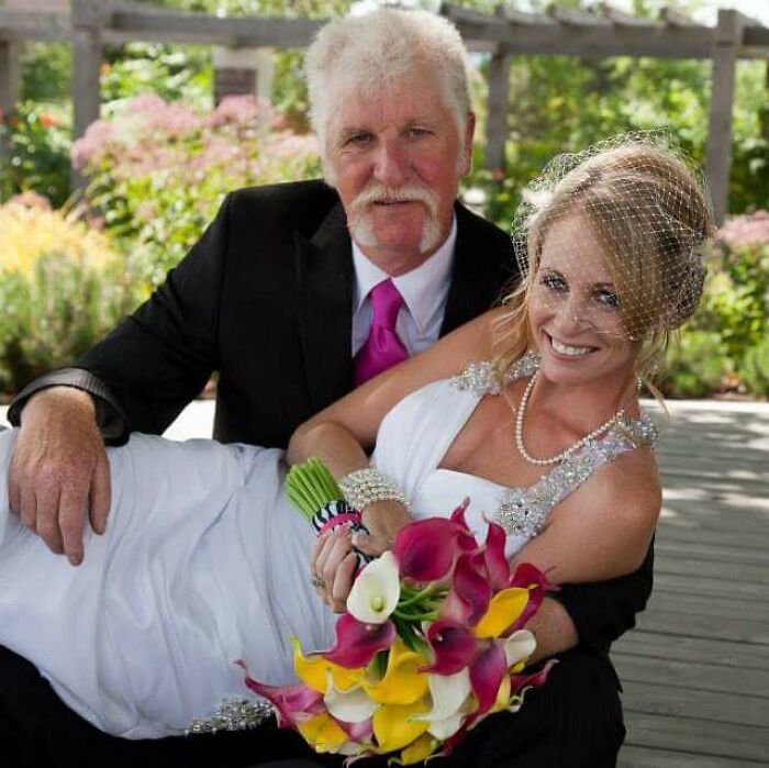 Bride in a wedding dress with birdcage veil holding a colorful bouquet, posing awkwardly with an older man in a suit outdoors.