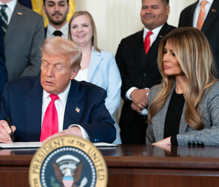 Melania Trump's body language observed closely as Donald Trump signs documents with onlookers behind them.