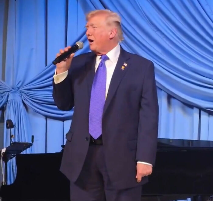Man in a suit holding a microphone on stage at a Mar-a-Lago event with blue draped curtains in the background.