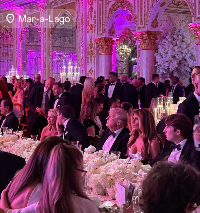 Guests dressed in formal attire at a lavish event inside Mar-a-Lago with elegant decor and pink lighting.