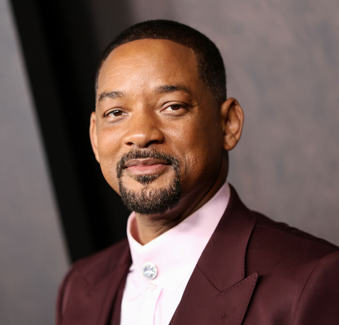 Will Smith wearing a maroon suit and light pink shirt, posing at an event related to a harassment lawsuit.