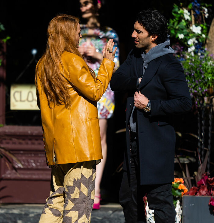 Blake Lively in a mustard jacket talking to a man outdoors, amid a scene drawing attention from fans and media.