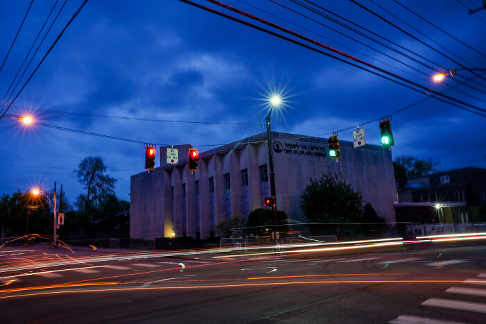 Building with Tree of Life memorial sign at dusk near a busy intersection with lit traffic lights and streaks of car lights.