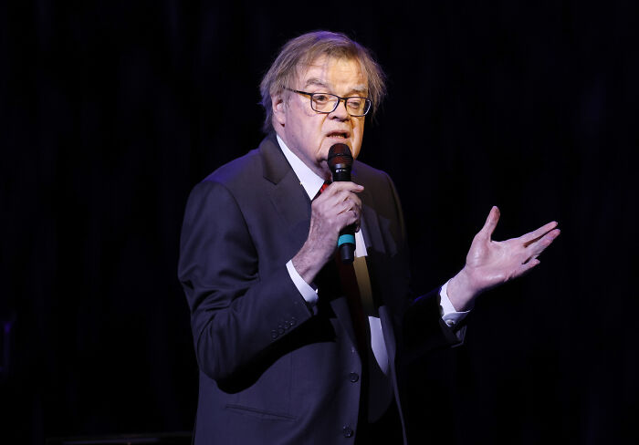 Older man in a suit holding a microphone, speaking on stage about celebrity downfalls in a dark background setting.