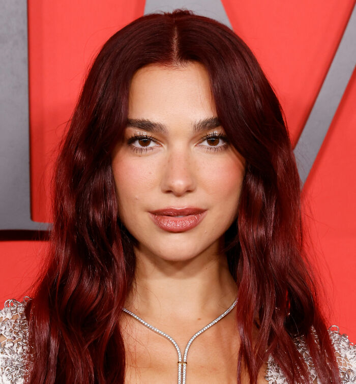 Dua Lipa posing with long red hair and subtle makeup at an event, wearing a silver necklace and shimmering outfit.