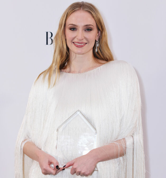 Sophie Turner smiling in a white outfit while holding an award, highlighting her Lara Croft transformation for Tomb Raider.