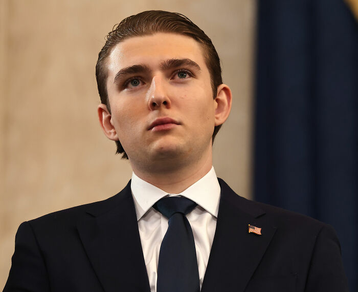 Young man in a dark suit and tie, wearing an American flag pin, looking serious against a neutral background.