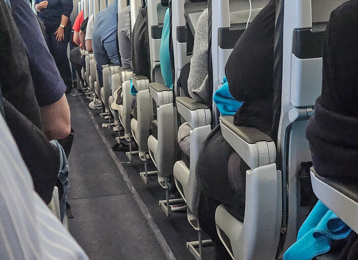 Cramped airplane cabin showing sardine can seating with narrow aisle and passengers squeezed into tight legroom.
