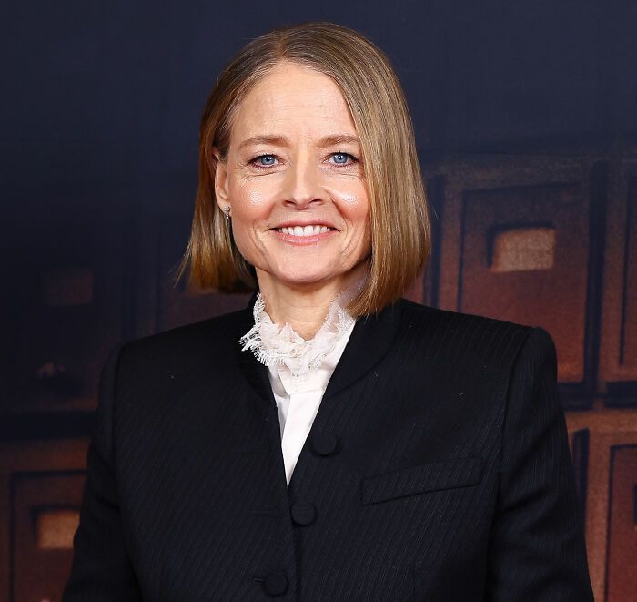 Jodie Foster smiling at an event, wearing a black suit with a white lace collar, highlighting Hollywood predators topic.