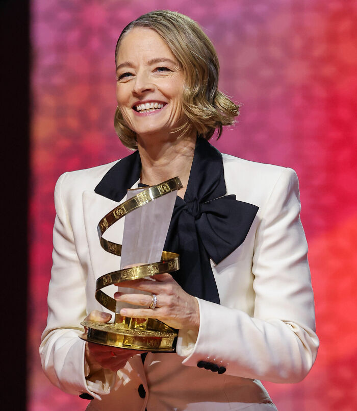 Jodie Foster smiling and holding an award, highlighting her story of being saved from Hollywood predators as a child.
