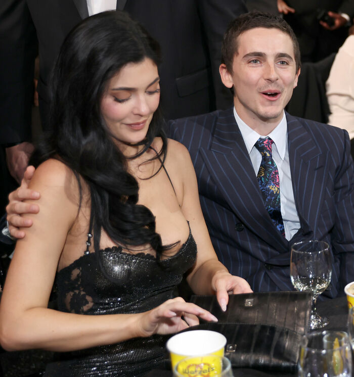 Timoth&eacute;e Chalamet seated beside Kylie Jenner at an event, dressed formally, engaging with the crowd around them