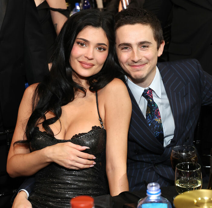 Timoth&eacute;e Chalamet smiling in a pinstripe suit next to a woman in a black dress at a formal event gathering.