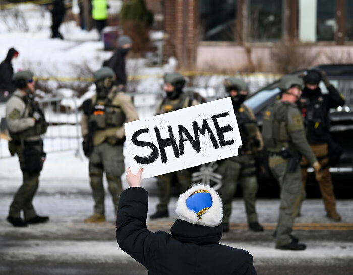 Protester holding a shame sign facing ICE agents in tactical gear during a public demonstration on a snowy street.