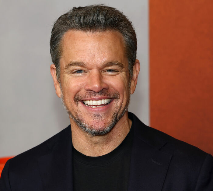 Matt Damon smiling in a dark jacket against a neutral background amid Amanda Knox cancel culture feud discussions.