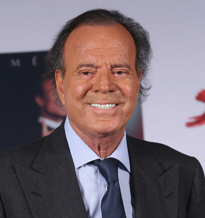 Julio Iglesias smiling at an event, dressed in a suit and tie, with news about investigation and accusations.