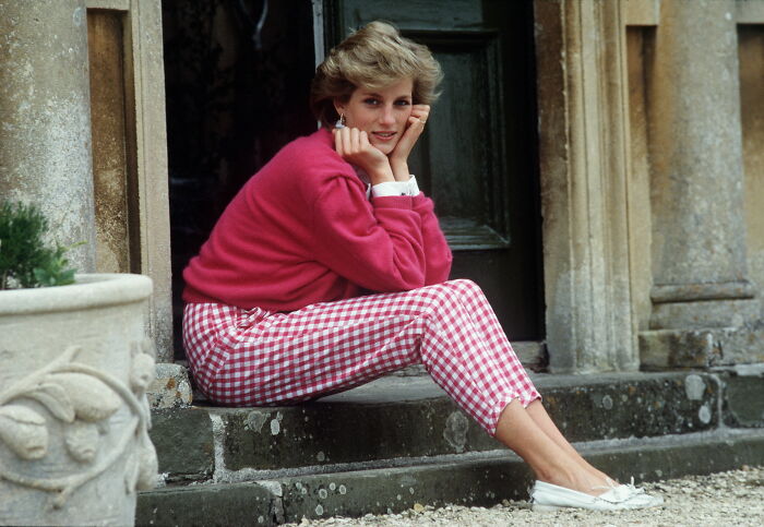 Woman sitting on stone steps outside a building, wearing a pink sweater and checkered pants, reflecting on celebrity downfalls.