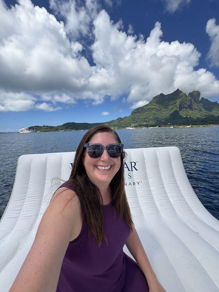 Woman wearing sunglasses smiling on a boat with blue water and French Polynesia mountains in the background.