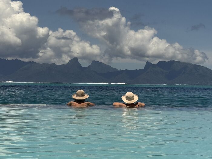 Two people wearing hats relax in a pool overlooking the vibrant blue waters of French Polynesia under cloudy skies.