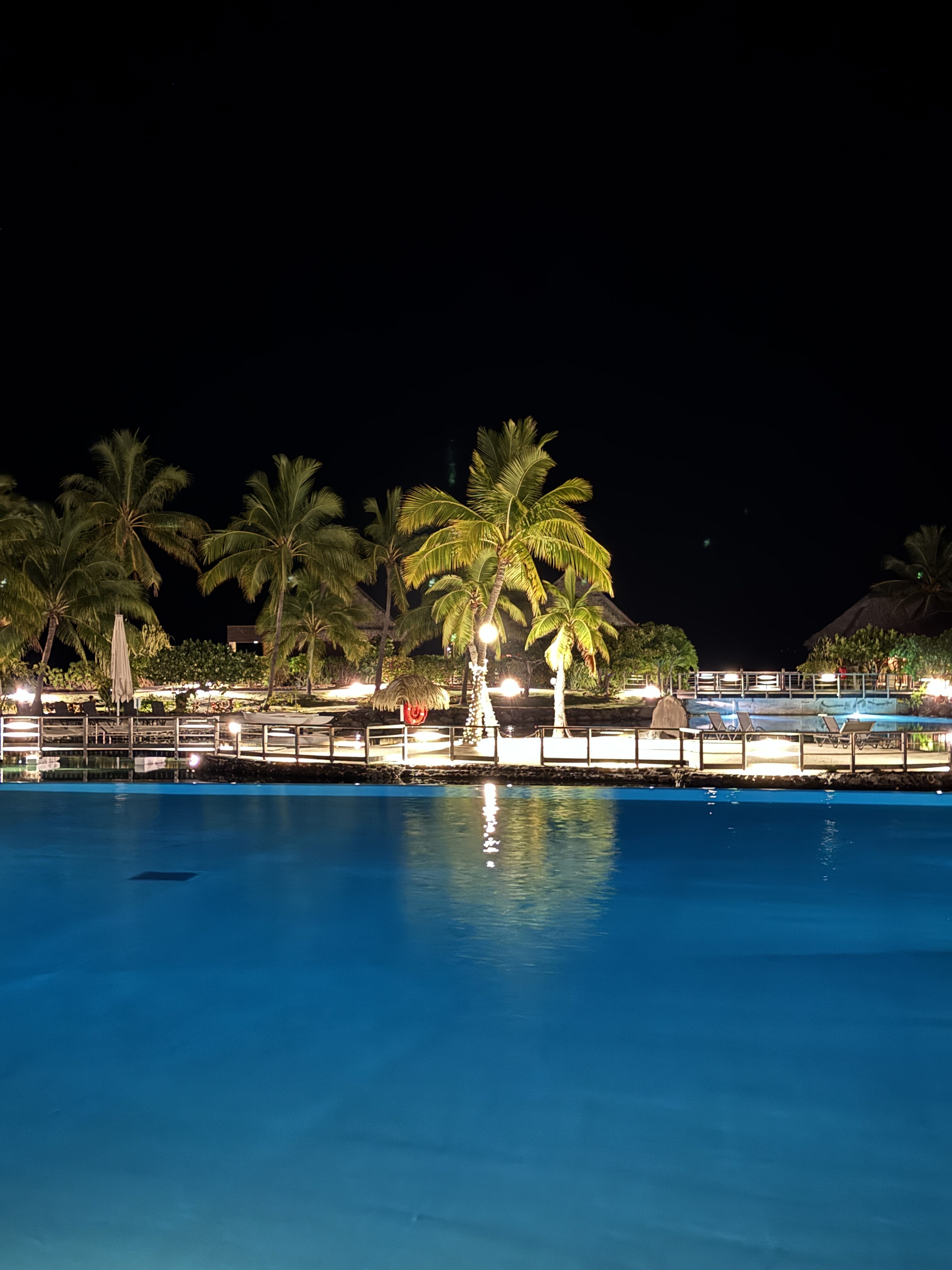 Nighttime pool with vibrant shades of blue water, palm trees, and lights reflecting in French Polynesia resort.
