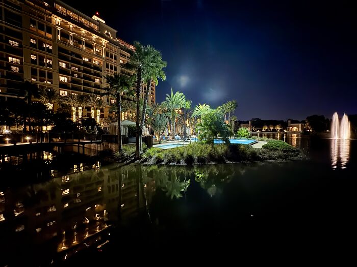 Night view of serene Central Florida resort with calm water, palm trees, fountains, and illuminated buildings reflecting beauty and silence.