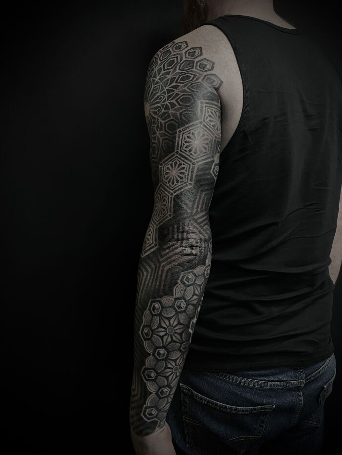 Intricate geometric tattoos covering full arm sleeve on person wearing black tank top and jeans against dark background