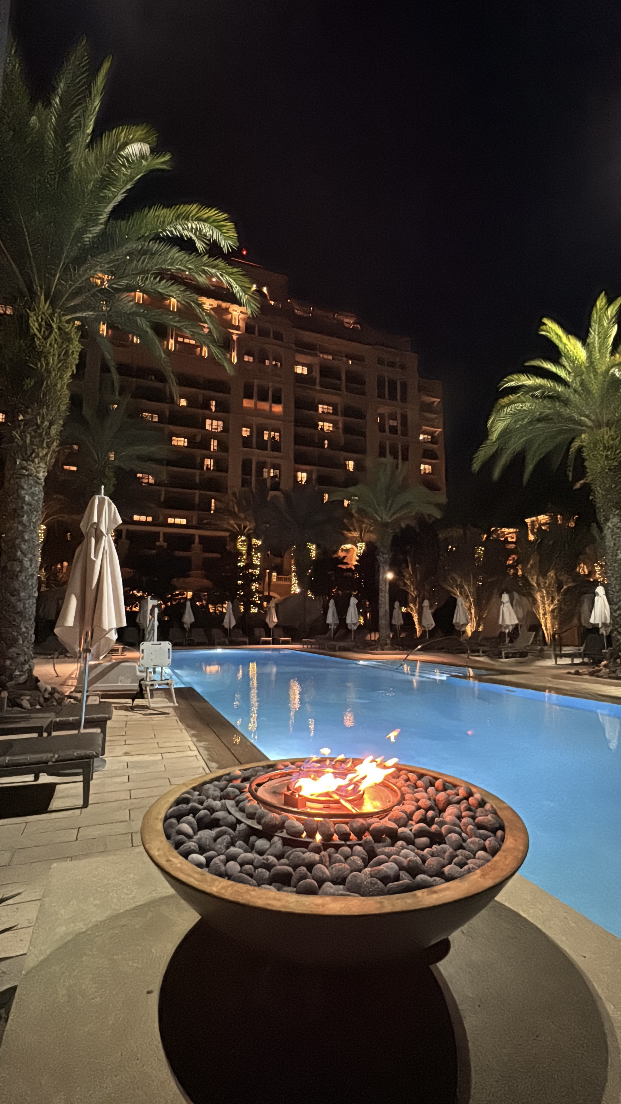 Nighttime view of a peaceful Central Florida pool area with fire pit, lit palm trees, and an illuminated hotel building.