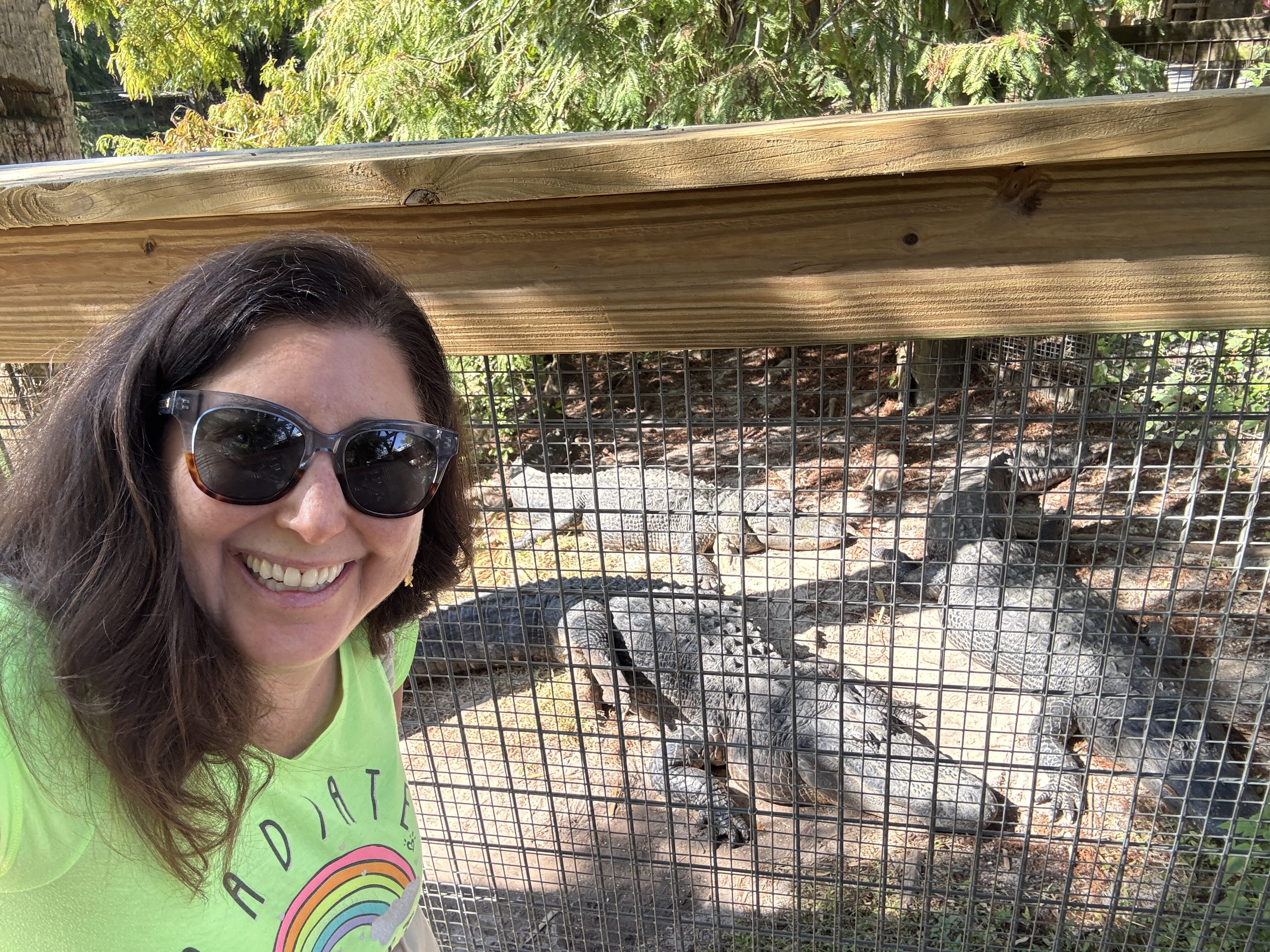 Woman wearing sunglasses and a bright shirt takes a selfie near fenced alligators in a quiet Central Florida setting.