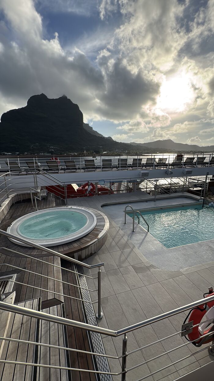 Cruise ship pool and hot tub with mountain and ocean views under dramatic blue skies in French Polynesia.