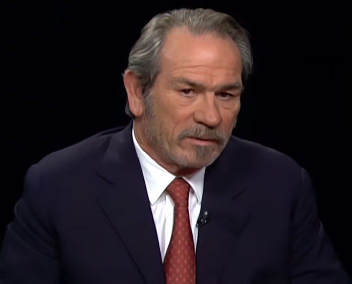 Actor Tommy Lee Jones in a dark suit and red tie during a serious interview on a black background.