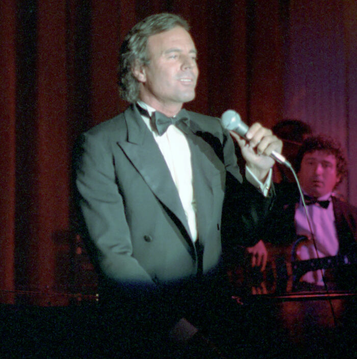 Julio Iglesias in a tuxedo holding a microphone on stage, related to Enrique Iglesias' famous father investigation news.