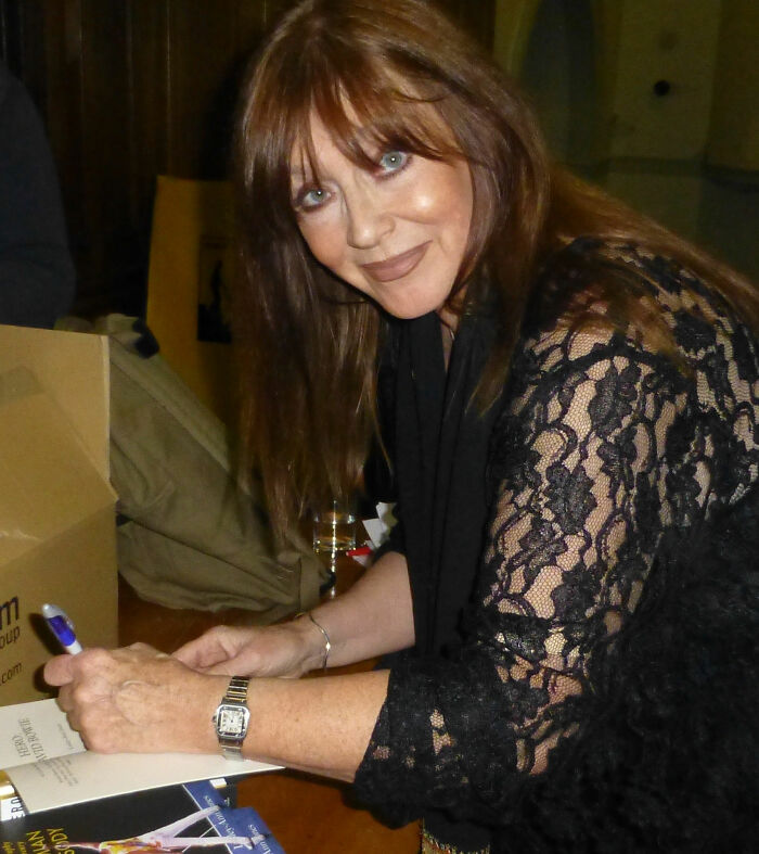 Woman with brown hair signing books at a table, related to Freddie Mercury's secret daughter news and recent passing at 48.